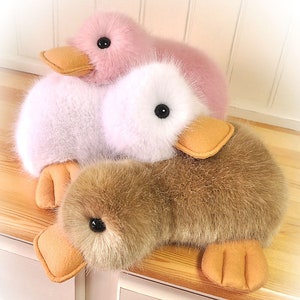 Peut inclure: Deux canetons en peluche, l'un rose et blanc et l'autre brun, avec des becs et des pattes orange. Les canetons sont couchés sur une surface en bois.