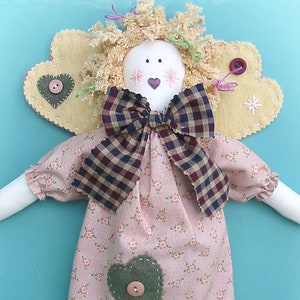 Peut inclure: Une poupée de chiffon ange faite à la main portant une robe rose à fleurs, un nœud en vichy brun et blanc et un cœur vert avec un bouton sur le devant. L'ange a une auréole en feutre jaune avec un bouton rouge et une étoile blanche.