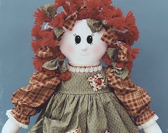 Könnte beinhalten: Eine handgemachte Stoffpuppe mit roten Wollhaaren, einem grün-braunen karierten Kleid und weißer Spitzenborte. Die Puppe hat ein weißes Gesicht mit gemalten Augen und rotem Rouge.