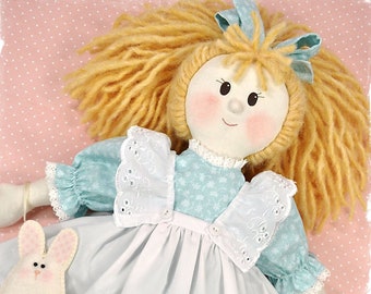 Könnte beinhalten: Eine handgefertigte Puppe mit blonden Haaren, die ein blaues und weißes Kleid mit einer weißen Schürze und einer weißen Spitzenborte trägt. Die Puppe hält eine kleine Hasentasche mit einem floralen Herzdesign.