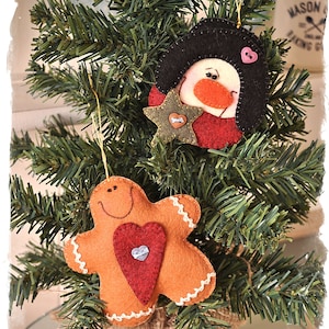 Könnte beinhalten: Zwei Filz-Weihnachtsbaumschmuckstücke hängen an einem kleinen künstlichen Weihnachtsbaum. Ein Ornament ist ein Lebkuchenmann mit einem roten Herzen und das andere ist ein Schneemann mit einem roten Hut und einem Stern.