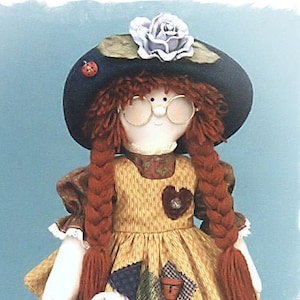 Peut inclure: Une poupée de chiffon faite à la main avec des cheveux roux, portant une robe jaune et marron, un chapeau noir avec une fleur et des lunettes. La poupée tient un lapin en peluche blanc avec des accents roses.