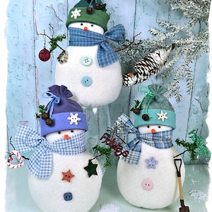 May include: Three handmade felt snowman ornaments with different colored hats and scarves. The snowmen are decorated with buttons and other embellishments. The snowmen are sitting on a blue background with snowflakes.