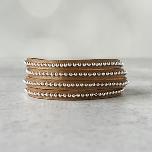May include: A wrap bracelet featuring multiple strands of tan leather. Each strand is adorned with a row of small, silver-colored beads. The bracelet is designed to wrap around the wrist multiple times, creating a layered look.