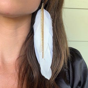 May include: A long, white feather earring with a gold-colored chain of small, sparkling stones. The earring is worn near the ear, with the white feather and chain hanging down. The background is blurred, with a glimpse of a light green wall.