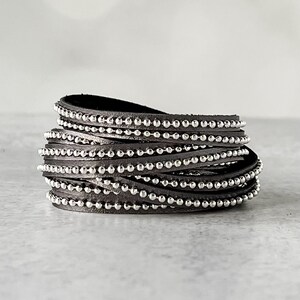 May include: A gray leather wrap bracelet with silver beads. The bracelet is made of multiple strands of leather that are wrapped around the wrist. The beads are evenly spaced along the leather strands.