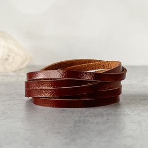 May include: A stack of dark brown leather strips, possibly for crafting or jewelry making, is displayed against a light gray background. The leather appears to be smooth and has a slight sheen, suggesting a polished finish. The strips are neatly arranged.
