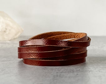 May include: A stack of dark brown leather strips, possibly for crafting or jewelry making, is displayed against a light gray background. The leather appears to be smooth and has a slight sheen, suggesting a polished finish. The strips are neatly arranged.