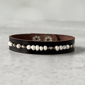 May include: A black leather bracelet with a row of white pearl-like beads and silver accents.