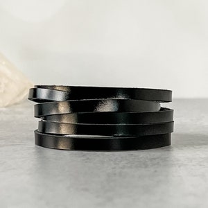 May include: A stack of six black leather strips, each approximately 0.25 inches wide, arranged in a slightly offset manner. The leather has a glossy finish, reflecting light. The strips are stacked on a light gray surface.