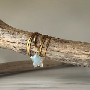 May include: A gold-toned necklace with a star pendant. The star has a light blue, iridescent center and a gold-colored border. The necklace features multiple strands of gold-colored, flat cord, wrapped around a piece of driftwood.