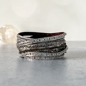 May include: A multi-strand bracelet featuring a dark brown leather band base. The bracelet is adorned with layers of silver-toned chains and sparkling rhinestones. The design is a mix of textures and metallic finishes, creating a statement piece.