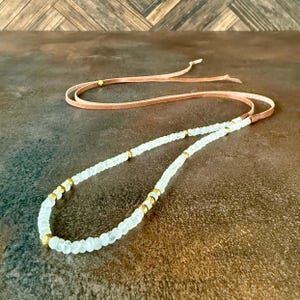 May include: A white beaded necklace with gold accents on a brown leather cord. The necklace is on a brown surface.