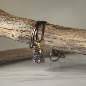 May include: A purple amethyst crystal pendant with iridescent accents hangs from a brown leather cord. The pendant is set in gold.