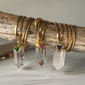 May include: Three gold necklaces with clear quartz crystal pendants. Each pendant has a small triangle-shaped gemstone at the top. The gemstones are green, pink, and purple.