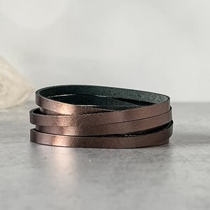 May include: A multi-layered leather bracelet in a rose gold color. The bracelet is made of thin strips of leather that are woven together.