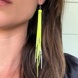 May include: Long, neon yellow fringe earrings with silver accents. The earrings have a simple hook design and feature multiple strands of bright yellow suede-like material, interspersed with silver-colored metal bars.