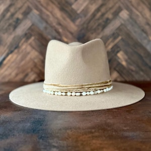 May include: A beige felt fedora hat with a wide brim and a band of white pearls and brown leather.