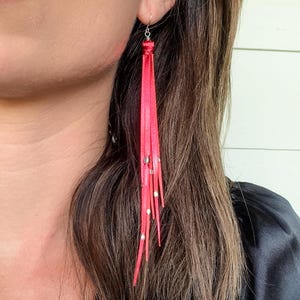 May include: A pair of long, red tassel earrings with silver beads. The earrings are made of leather and have a fringe design.