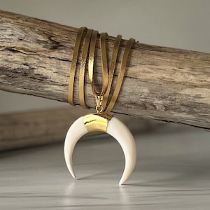 May include: A gold and white crescent moon pendant necklace with a brown leather cord. The cord is wrapped around a piece of driftwood.