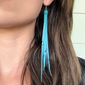 May include: Turquoise leather fringe earrings with silver-tone metal accents. The earrings feature long, thin leather strips gathered at the top and adorned with small, metallic studs. The earrings are designed to dangle and sway.