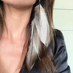 May include: Long, dangling feather earrings. The earrings feature multiple feathers in shades of white, gray, and brown. The feathers are attached to a delicate silver chain. The earrings are a bohemian style.