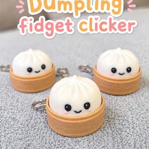 Puede incluir: Tres fidget clickers de dumpling blancos, cada uno con una cara sonriente de ojos negros y mejillas rosadas, colocados en una base marrón estilo vaporera de bambú con un llavero de metal. El texto "Dumpling fidget clicker" aparece arriba.