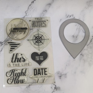 May include: A clear sheet of stamps with black ink featuring phrases like "Enjoy the Journey", "Field Notes", "This is the Life", "Right Here For You", and "Right Now". There is also a compass and a date stamp.