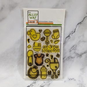 May include: Clear photopolymer stamps featuring baby-themed designs, including a rubber duck, rattle, baby bottle, and stroller. The text "we grew by two feet" is also included. The stamps are packaged in a clear plastic sleeve.