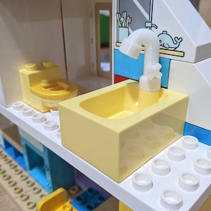 May include: A white plastic LEGO faucet with a yellow bathtub and a white toilet in a white and blue LEGO bathroom set.