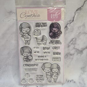 May include: A clear stamp set featuring illustrations of pregnant figures, baby-related images, and text. Text includes phrases like "Rock That Baby Bump!", "It's a Boy!", and "Congrats!" Designed in France.