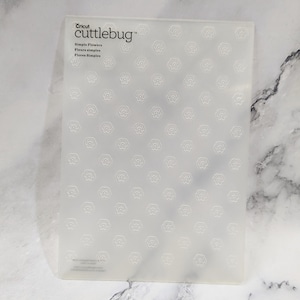 May include: A clear plastic sheet with a repeating pattern of small flower shapes. The sheet is labeled "Cricut cuttlebug" and "Simple Flowers, Fleurs simples, Flores Simples".