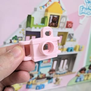 May include: A pink plastic toy camera with a lens and a handle. The camera is a plaything for children.