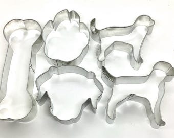 May include: A collection of silver-colored metal cookie cutters in various shapes, including a dog bone, paw print, and two dog silhouettes. These cutters are designed for making dog-themed treats.