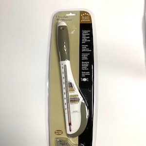 May include: A Taylor Connoisseur Series chocolate thermometer in a clear plastic package. The thermometer has a grey handle and a white face with temperature markings in Fahrenheit. The thermometer is designed for chocolate making.