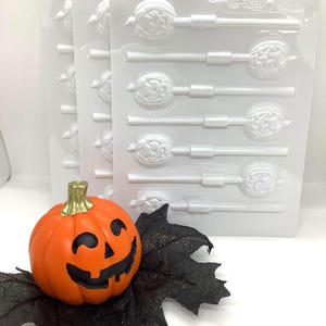 May include: An orange pumpkin with a smiling face and a gold stem rests on black glitter leaves. Behind it are clear plastic lollipop moulds with Halloween designs, ideal for making themed sweets.