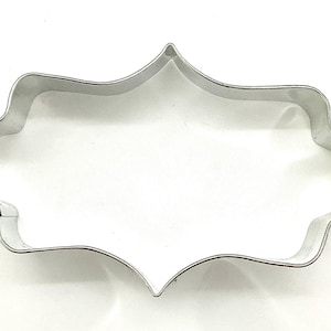 May include: A silver-colored metal cookie cutter in a decorative, ornate shape. The cutter has a scalloped edge and a pointed top and bottom. The shape is reminiscent of a decorative plaque or emblem.
