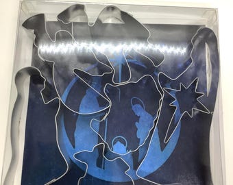 May include: A collection of silver metal cookie cutters in diverse shapes, such as a star, camel, and figures, presented against a blue and white backdrop. The cutters are packaged in a clear plastic box, possibly for a festive or religious occasion.