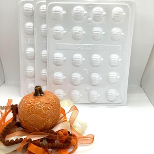 May include: Three white plastic pumpkin-shaped candy molds with the text "SH-3004 CK". An orange ceramic pumpkin with leaf detailing sits near orange and cream-colored ribbons and trim. A festive autumn-themed image.