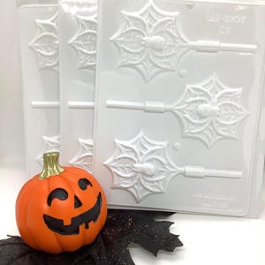 May include: Three clear plastic candy molds with a spiderweb design, perfect for Halloween treats. An orange pumpkin with a carved face sits in front of the molds. Black decorative leaves add to the festive theme.
