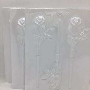 May include: Three white plastic molds for making rose-shaped lollipops. Each mold has a rose design with a stem and leaves. The molds are stacked, showing the front of each mold. The text "2H-3114 OK" is visible on one mold.