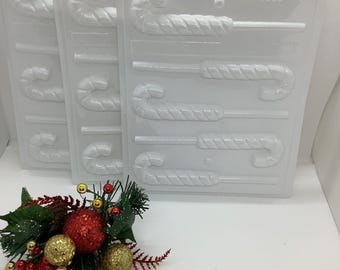 Puede incluir: Moldes de plástico transparente para bastones de caramelo. Los moldes tienen múltiples cavidades para crear bastones de caramelo. Un arreglo festivo de adornos y vegetación rojos y dorados brillantes está en primer plano. Los moldes están etiquetados "CA-4303".