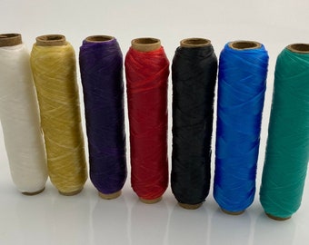 May include: Eight spools of thread in various colors, including white, yellow, purple, red, black, blue, and green. The spools are arranged in a row from left to right.