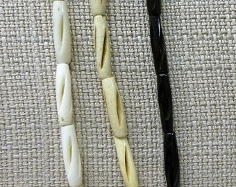 May include: Three strands of beads, two white and one black, with a carved design. The beads are strung on a cord and are hanging against a beige fabric background.