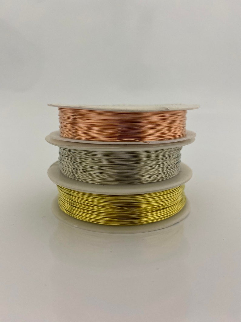 May include: Three spools of wire, one gold, one silver, and one copper, are stacked on top of each other. The spools are on a white background.