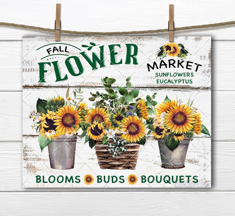 May include: A white wooden sign with the text "Fall Flower Market" in green and black lettering. The sign features three arrangements of sunflowers, eucalyptus, and other flowers in buckets and a basket. The text "Blooms Buds Bouquets" is at the bottom of the sign.