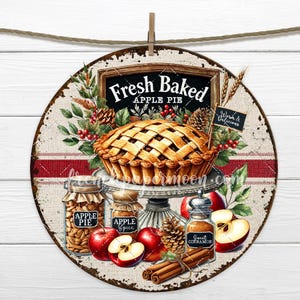 May include: A round, rustic-style sign with a red and white striped border. The sign features a freshly baked apple pie with a golden crust, surrounded by apples, cinnamon sticks, and jars labeled "Apple Pie" and "Apple Spice". The sign reads "Fresh Baked Apple Pie" in a black chalkboard style font.