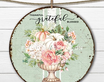 May include: A round, rustic metal sign with a distressed green background and white stripes. The sign features a floral arrangement of pink roses, white hydrangeas, and green leaves in a white urn. Two pumpkins, one white and one peach, are nestled in the arrangement. The sign reads "Thankful grateful Blessed".