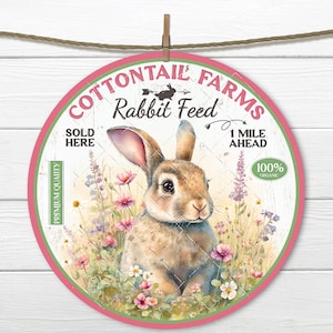 Peut inclure: Une pancarte ronde rose et verte avec le texte "Cottontail Farms Rabbit Feed" et un lapin de dessin animé dans un champ de fleurs. La pancarte indique également "Sold Here" et "1 Mile Ahead 100% Organic".