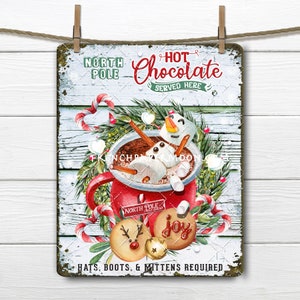 Puede incluir: Un letrero de metal de estilo vintage con una escena navideña festiva. Un muñeco de nieve está sumergido en una taza de chocolate caliente con malvaviscos. El letrero dice "Polo Norte Chocolate Caliente Servido Aquí" y "Sombreros, Botas y Mitones Requeridos".